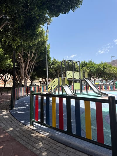 This is an outdoor photograph of a playground in Campillo Del Moro, Spain, on a sunny day. The playground features a modern slide structure with two metal slides, one steeper than the other, and a black metal frame with lime green panels. The playground floor is a soft, multi-colored surface. A colorful fence with red, yellow, blue, and black vertical bars surrounds the playground area. To the left of the playground, there is a paved walkway made of brick and concrete, shaded by mature trees with exposed roots and branches. In the background, more trees line the street, and buildings with red roofs and windows are visible. A street lamp and a few parked cars are also present. The sky is clear blue with a few wispy white clouds. No people are visible in the image.