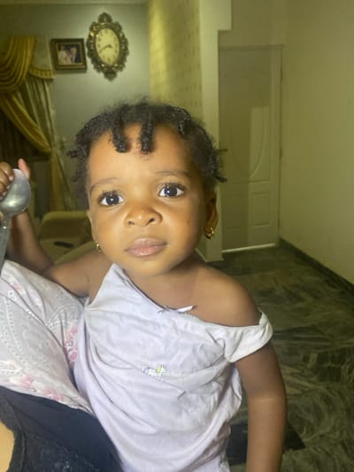 A young child with dark, curly hair and gold hoop earrings looks directly at the viewer. The child is wearing a light purple shirt adorned with a small unicorn. The child is holding a silver spoon. In the background, there is a white door, a clock, framed picture, and a curtain. The floor is marble.