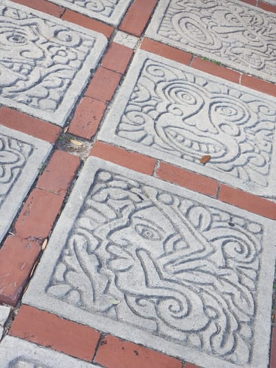 This is a close-up, high-angle shot of a paved pathway featuring decorative square concrete tiles interspersed with red bricks. The concrete tiles are intricately carved with abstract, stylized faces or masks, featuring swirling patterns and bold lines. The bricks are laid in a pattern that complements the concrete tiles. The pathway appears to be outdoors, possibly in a park or public space. The lighting suggests it is daytime, with sunlight casting shadows and highlighting the textures of the materials. There are a few fallen leaves and some sparse green vegetation visible in the crevices between the tiles and bricks. The overall impression is one of detailed craftsmanship and a possibly cultural or artistic design choice for the paving. The location in the city of Maitland, United States of America, is not directly evident from the image content itself, but the style of the carvings might suggest a specific cultural influence if known.