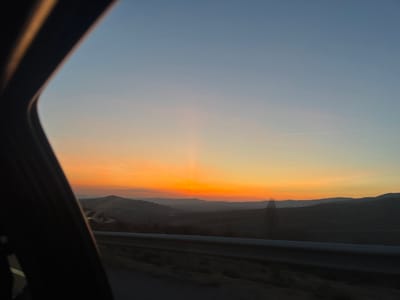 The image presents a sunset over a mountainous landscape near Casas del Puerto, Spain, viewed from the interior of a vehicle. The left third of the frame shows the dark, curved structure of the vehicle's interior. The sky exhibits a clear gradient, transitioning from intense orange and yellow hues at the horizon to light blue in the upper portion, with a faint vertical light pillar rising from the brightest orange band. A thin, linear contrail is visible across the upper-middle sky. In the foreground, a dark grey metal guardrail runs horizontally, indicating a road. Beyond the guardrail, a series of dark, undulating hills and mountains are silhouetted against the colorful sky, showing indistinct vegetation. No individuals or specific actions are discernible within the scene.