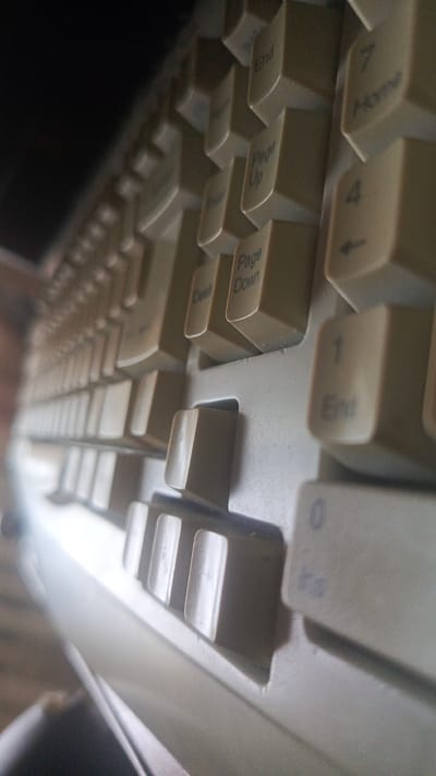 This is a close-up, low-angle shot of a beige computer keyboard. The perspective emphasizes the depth of the keys and the rows of the keyboard, with a dark, out-of-focus area on the left, possibly a monitor or a dark background. The keys visible include "End", "Home", "Page Up", "Page Down", "Delete", "1", and "0". Some keys show faint lettering, while others are mostly blank. The lighting suggests an indoor setting, possibly with artificial light. The keyboard appears to be older, given its color and design. No people or specific activities are depicted, just the object itself. The location context provided is Kardonikskaya, Russia, but there are no visual cues in the image to confirm this. The overall mood is neutral, focusing on the texture and form of the keyboard.