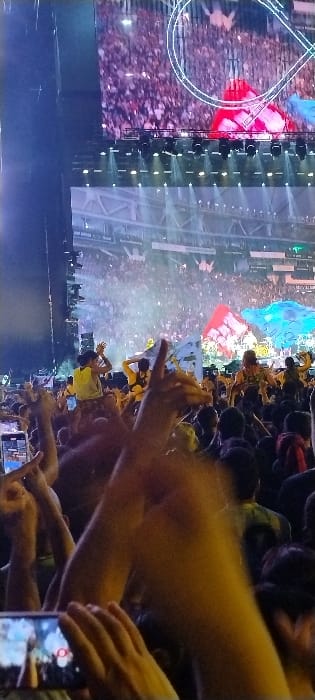 This image captures an energetic night-time concert or festival scene, viewed from within a large crowd. The foreground is filled with numerous attendees, many with their arms raised in excitement. Several individuals are holding up mobile phones, seemingly recording the event or capturing photos.
In the mid-ground and background, a large, brightly lit stage is visible, featuring multiple spotlights casting beams of light upwards and across the performance area. A massive video screen behind the stage displays dynamic graphics, including a prominent circular outline. The crowd extends far back from the stage, suggesting a large-scale event. Among the raised hands, several colorful flags are visible, notably a red and white one, and a blue and white one, contributing to the festive atmosphere.
The setting is clearly illuminated by artificial light, indicating that the event is taking place at night. The overall impression is one of enthusiasm and active engagement from the audience. The location context provided is Tolosa, Argentina, suggesting a lively cultural event in that area. No discernible text is visible on any flags, screens, or other objects within the image.