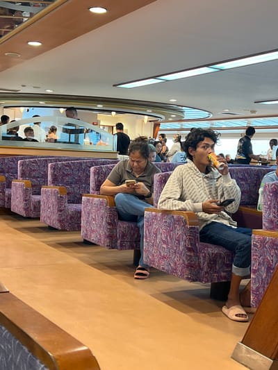 This image depicts the interior of a ferry or train with rows of upholstered seating. The seats have a distinctive purple and pink patterned fabric. Several individuals are seated, with the primary focus on a young man in the foreground wearing a white hooded sweatshirt with "NEW YORK" repeatedly printed on it. He is holding a mobile phone in one hand and a beverage in the other, appearing to drink from it. Another person is seated next to him, looking at their phone. The overall lighting suggests daytime, likely indoor ambient light supplemented by natural light from windows that are not fully visible. The scene appears to be a public transportation setting, possibly during a journey. Visible text includes repeated instances of "NEW YORK" on the man's clothing and "AED" on a sign in the background, likely indicating an Automated External Defibrillator. The location context is Don Sak, Thailand, which suggests this could be a ferry operating in that region.