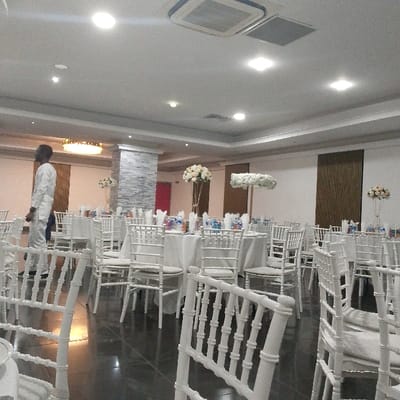 This image depicts an indoor event space, identified as being within the city of Birmingham, United Kingdom. The scene shows a banquet hall or reception room fully prepared for an event, featuring numerous circular tables draped with white tablecloths and surrounded by white Chiavari-style chairs, some with white seat cushions. Table settings include bottled beverages and what appear to be red-folded napkins. Tall gold stands with floral arrangements of white, cream, and light pink flowers serve as centerpieces, alongside a distinct, elongated horizontal arrangement of white flowers suspended above a central table.
The room possesses a light-colored ceiling with recessed lighting and visible air conditioning units. The floor is dark and reflective. A central pillar is faced with grey, stacked stone-like material, with a red door partially visible to its left. Dark wood paneling adorns a wall section on the right, and a chandelier is visible on the far-left wall.
One individual stands on the far-left, facing away from the camera and looking right. This person wears a light-colored, two-piece traditional garment. No other individuals or active interactions are observed within the prepared environment.