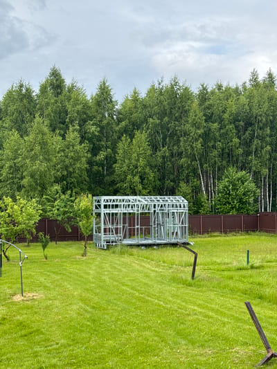 An exterior, eye-level, medium shot captures a partially constructed metal frame for a greenhouse or small building situated on a trailer in a grassy, rural yard. The structure is metallic gray, consisting of a grid of interconnected beams and supports, with an open framework suggesting it is in the early stages of assembly. Behind the frame, a brown wooden fence stretches across the background, separating the yard from a dense forest of green deciduous trees, primarily birch trees with their characteristic white bark. The sky above is overcast with gray clouds. The foreground and midground are covered in well-maintained, vibrant green grass. Scattered across the lawn are a few young trees and some metal stakes, one of which has a piece of equipment attached. The overall scene suggests a residential property undergoing construction or development in a natural, possibly remote, setting. The image is taken during daylight hours.
