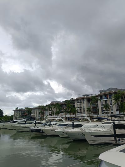 This image depicts a row of luxury yachts docked in a marina in Ko Kaeo, Thailand. The marina is situated in front of a row of modern buildings with balconies and palm trees. The sky is overcast with dramatic, heavy clouds, suggesting impending rain or stormy weather. The water in the marina is calm, reflecting the yachts and the buildings. No people are clearly visible in the image.
