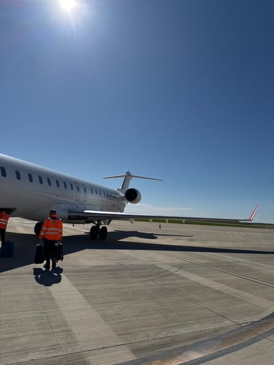 The image depicts an airport scene in Alguaire, Spain. A white passenger jet, tail number EC-NLC, is parked on the tarmac. Two individuals, dressed in orange safety vests, appear to be loading luggage. Shadows are cast by the plane and people, indicating bright sunlight. The sky is clear and blue. The airport runway surface is visible with concrete markings and a distant green area on the horizon. The tail of the jet has a red and white design.