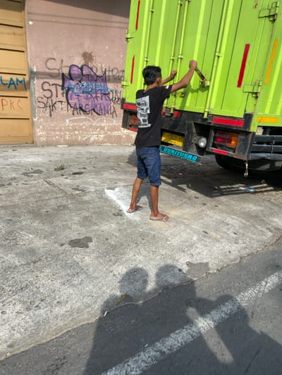 The image shows a man standing beside a green truck in Purwakarta, Indonesia, at coordinates -6.558, 107.437. The man is wearing a black T-shirt and blue jeans and is facing the truck. He is reaching his arm up to grab a handle on the truck. The truck is parked on the side of the road and is facing away from the camera. The environment is a paved road with a painted white stripe on the right side. The road is partially shaded by the truck. The man is standing in front of a light-colored wall with graffiti. There is a shadow of the man on the ground. The truck has a black bumper and a license plate that says "3824 TI". There is a sticker on the truck that says "S. UBERIYERD" in blue letters.