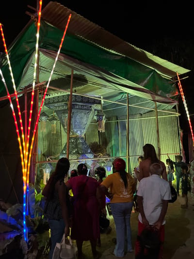 The image depicts an outdoor scene at night, likely a market or a gathering, in Habaraduwa, Sri Lanka. Several people are visible, some standing and others seated or leaning. They are gathered around a temporary structure covered with green tarp and corrugated metal sheeting, which appears to be a stall or a covered area.
The scene is illuminated by strings of colorful LED lights, predominantly red, yellow, and white, which are draped around the structure and extending outwards. These lights create a festive atmosphere. Several lanterns, decorated with intricate patterns in dark blue and white, are hanging within the structure, suggesting a possible cultural event or decoration. Some of the people are wearing casual clothing, and one person is wearing a red helmet.
The ground appears to be unpaved, consistent with an outdoor setting in a tropical location. The overall impression is of a lively nighttime event, possibly a festival or a local market, characterized by the vibrant lighting and the presence of decorative lanterns. The time of day is clearly night, indicated by the darkness surrounding the illuminated area. There is no visible text in the image.