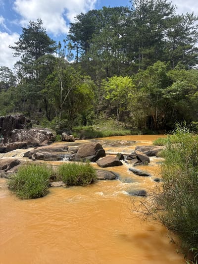 The image depicts a section of a river in Păng Tiêng 1, Vietnam. The water is a murky brown-yellow color. The river flows over and around dark-colored rocks and boulders. Green vegetation grows along the riverbanks. Trees are present on one side of the river, while the other side shows rocks and a cloudy blue sky.