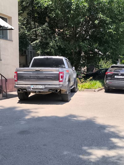 A silver Ford F-150 Raptor pickup truck is parked in front of a building, with a black Toyota RAV4 parked to its right. The Ford truck has a license plate that reads "KZ 826 BRZ 02". The Toyota RAV4 has a license plate that reads "KZ 099TAH 02". The scene appears to be a residential area or a parking lot, with trees providing shade. The image is taken during the daytime, under bright sunlight, casting shadows on the ground. The building to the left is light-colored with a window and an awning. The overall environment is outdoors, with pavement and vegetation visible.