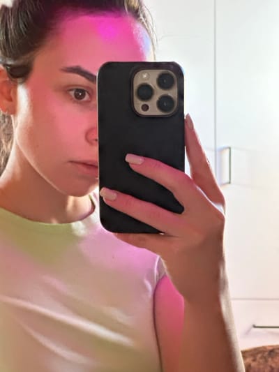 An indoor mirror selfie depicts an adult female subject in the city of 3-Y Uchastok, Russia. The subject's face, partially obscured, is illuminated from the left by a strong pink/magenta light source, casting a noticeable hue across her forehead and the visible right side of her face. Her right eye is open, looking directly towards the mirror. She has dark hair and fair skin. Her right hand, with fingernails painted a light neutral color, holds a dark-colored smartphone, equipped with a triple camera system, positioned centrally in front of her face. She is dressed in a light-colored, likely light green, crew-neck t-shirt. The background is a plain white surface, possibly a wall or wardrobe, with a subtle handle visible on the right side.