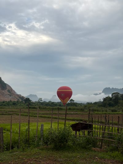 An outdoor, medium shot captures a hot air balloon in the sky above a rural landscape. The balloon is predominantly red with yellow accents and a flag displaying a white emblem against a red background. It is positioned slightly to the left of the frame's center. The surrounding environment includes tiered rice fields, a bamboo fence, and lush green foliage. In the background, rolling hills and mountains are visible, partially obscured by clouds. The sky is overcast with a mix of gray and white clouds. The image appears to be taken in Hong Ngern Village, Laos. No people are visible in the frame.