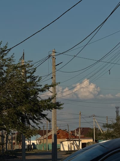 The image shows a residential area with power lines crisscrossing the sky. Several utility poles are visible, supporting a complex network of wires. In the background, houses with various roofing materials, including red and brown, are clustered together. A green fence and some trees are also present, adding to the rural or semi-rural feel of the scene.

The lighting suggests it might be late afternoon or early evening, as the sky is a clear blue with a few wispy clouds, and there is a warm glow to the scene. The overall impression is one of a quiet, everyday setting. There is no text visible in the image. The orientation of the image is such that the ground is on the right side and the sky is on the left, indicating the photo was taken on its side.