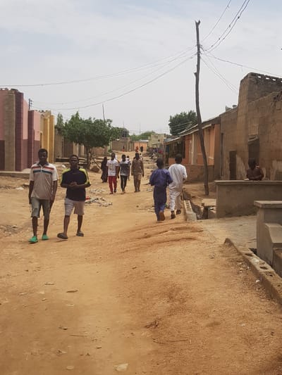 A street in Dutsinma B, Nigeria, shows a group of people walking along a dirt road flanked by buildings. The building on the left is a pinkish hue, and another building is on the right. Overhead, an unadorned utility pole with wires. Two men stand in the foreground. The first one is wearing a white shirt with a blue stripe and gray shorts. The second one is wearing a black shirt with yellow stripes and gray shorts.