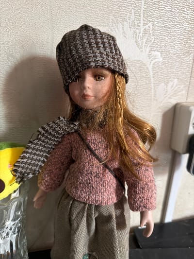 A doll with auburn hair, wearing a patterned cap, a pink knitted sweater, a plaid scarf, and a checkered skirt stands against a textured wall. A yellow object is partially visible to the left, next to a clear glass vase. To the right, a white electrical outlet is mounted on the wall. The scene appears to be an indoor setting, possibly a home. No specific location in the city of Shepsi, Russia is indicated in the image.