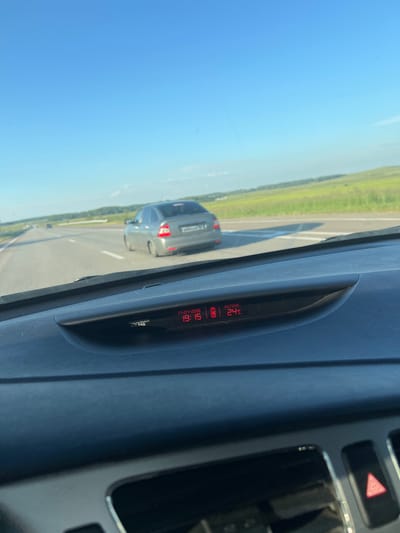 A gray sedan is driving on a highway in Burannaya, Russia. The sky is blue with a few clouds. The road is paved with white lines. There are fields of green grass and trees on either side of the road. The car's interior is visible, showing a dashboard with a digital display that reads "OUTSIDE 24C". The date is also displayed as "17-07-2026". The car is in motion, as indicated by the blurred background.
