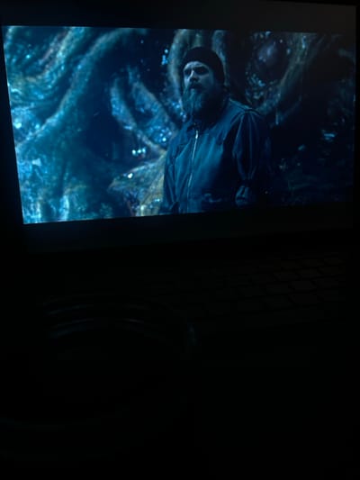 A photograph captures a screen displaying a dark, blue-toned scene featuring a male subject and an abstract background. The man, seen from the chest up, has a prominent dark beard and mustache, wears a dark beanie, and a dark zippered jacket. His gaze is directed upward and to the viewer's left. Behind him, the background consists of swirling, textured, dark blue and teal forms, possibly organic or crystalline, with scattered small, bright, shimmering light points. The bottom half of the photograph is almost entirely black, revealing only faint outlines of a keyboard's keys in the lower right, indicating the image was captured in a darkened room, likely from the perspective of someone viewing the screen (possibly a laptop or projected image). No specific location within Kernen im Remstal, Germany, can be identified from the visual content.