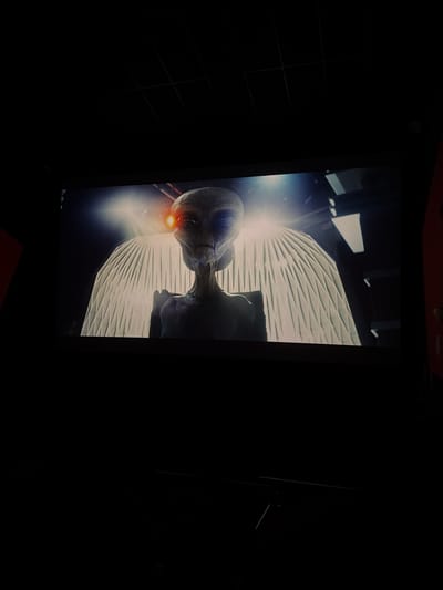 The image shows a close-up of a movie screen in a dark theater. The screen displays a digital rendering of an alien with large, dark eyes and a reddish glow emanating from its left eye. The alien has a gaunt, humanoid face and appears to have wing-like structures behind it, which are illuminated by a bright white light. The screen is framed by the dark interior of the theater, with the ceiling visible at the top and the edge of a red seat on the right side of the frame. The overall scene suggests a cinematic viewing experience, likely in a public cinema. The location of Ondara, Spain, is not depicted in the image.