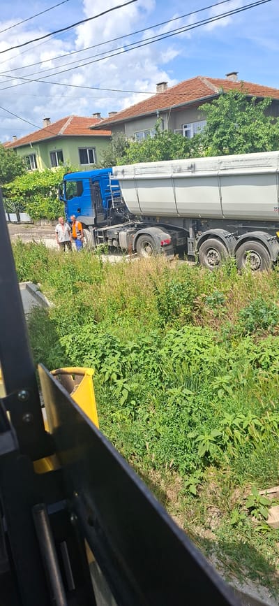 In Strazhitsa, Bulgaria, two men stand on a dirt road next to a blue semi-trailer truck with a grey hopper trailer. The man on the left wears a striped shirt and dark pants. The man on the right wears an orange vest over a blue shirt and blue pants. The truck and trailer are parked on the side of the road, which is lined with tall green grass and some wild plants. Behind the truck, a row of houses with red-tiled roofs and green facades is visible, surrounded by trees and foliage. The sky is partly cloudy. The foreground shows a portion of a yellow and black object, possibly the front of a vehicle or construction equipment.
