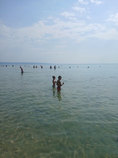 The image depicts a seaside scene with several people in the water. Two children are prominently featured in the foreground, with one boy in red shorts and another in blue shorts. They appear to be enjoying themselves in the shallow water. In the background, numerous other individuals are scattered across the water, some appearing to swim or wade. The water is clear and calm, with the sandy or pebbled bottom visible in some areas, suggesting a beach or shallow coastal environment. The sky is bright and partially cloudy, indicating it is likely daytime. The overall atmosphere suggests a relaxed and pleasant day at the beach, possibly during summer. The location context of Kranevo, Bulgaria, suggests this is a Black Sea beach. No visible text is present in the image.