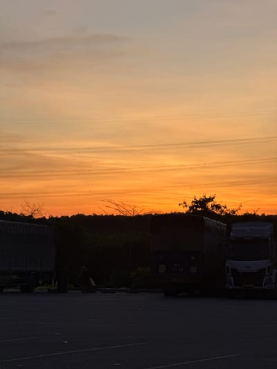 The image depicts a sunset over a truck parking area. The sky is a gradient of orange and yellow, with thin, wispy clouds. Several power lines stretch horizontally across the sky. In the foreground, the silhouettes of two large trucks are visible. The truck on the left is partially covered with a tarp, while the truck on the right is a modern tractor-trailer. The ground appears to be asphalt, marked with white lines. A dark, bushy treeline is visible behind the trucks, suggesting a natural or semi-rural environment. The lighting suggests it is either dawn or dusk. No specific landmarks or features place this scene within the city of Huế, Vietnam.