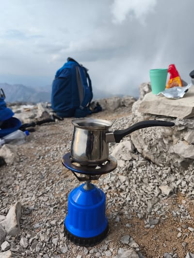 A small, shiny metal pot, likely a cezve, sits atop a portable blue gas stove, actively heating a liquid, on a rocky and sandy ground surface. The stove's burner shows metallic discoloration from heat. In the immediate background to the left, a crumpled clear plastic bottle and a portion of blue fabric are visible. Further back, a dark blue hiking backpack rests on the ground. To the right, a light green plastic cup and an open foil packet with red packaging rest on a flat, light-colored rock formation. The environment is an uneven, rocky, high-altitude terrain, with distant mountains partially obscured by a heavily overcast, cloudy sky. The scene is located in the city of Mali Mikulići, Montenegro, indicating an outdoor, potentially high-altitude, cooking or rest stop. No individuals are present.