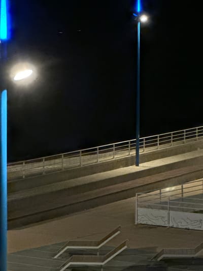 The image depicts an outdoor scene at night in Saint-Valery-en-Caux, France, likely a waterfront or promenade area. In the foreground, there are tiered seating areas with wooden benches, illuminated by artificial lights. A blue lamppost is visible on the left side of the image, with a bright light source at its top. Further back, a ramp or walkway with a white railing ascends into the darkness. Another blue lamppost stands higher up, also with a light source. The sky is dark and clear. The overall impression is a modern, illuminated public space.