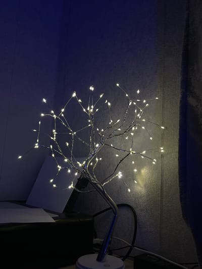 An artificial LED light tree lamp with a metallic trunk and branches, adorned with numerous small, white lights, is positioned on a desk. The background consists of a dark blue wall and a textured grey wall, with a shadow cast by a curtain on the right side. In the foreground, to the left of the lamp, is a black printer with a sheet of paper on it. Various cables and a power strip are visible on the desk near the lamp's base. The overall lighting is dim, with the primary illumination coming from the lamp itself. The scene is indoors, on a surface that appears to be a desk or tabletop.