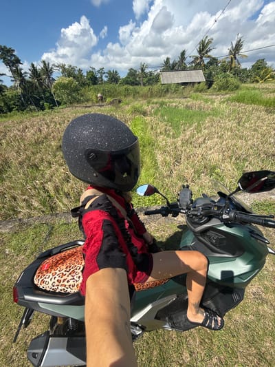 A woman wearing a black and glitter helmet and a red and black lace outfit is straddling a green motorbike on a grassy field. The woman's left leg is visible, showing a tattoo on her ankle, and she is wearing black sandals. Behind her is a brown animal print bag. The motorbike is parked on green grass with a view of a blue sky with white clouds, trees, and a small wooden structure. The location is identified as being in Mas, Indonesia, based on the visual characteristics.