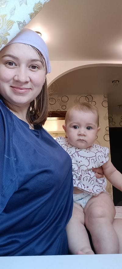 A woman wearing a light purple headscarf and a dark blue shirt is holding a baby. The baby is wearing a white onesie with purple bunny outlines and a diaper. The woman is looking towards the camera with a slight smile, while the baby is looking slightly off-camera with a neutral expression. They are indoors, with a textured wallpaper featuring a circular pattern in the background. The location is identified as Хохлы, Russia.