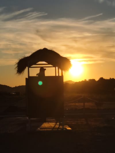 A silhouetted figure, possibly a person, stands within a thatched-roof structure at sunset. The structure appears to be a small, elevated booth or observation post. The sun is low on the horizon, casting a warm, orange glow across the sky and illuminating thin, wispy clouds. The foreground is dark and indistinct, with hints of ground and possibly a wooden platform. In the background, a silhouette of trees and hills is visible against the bright sky. The overall scene suggests a tranquil, natural setting. The specific location is Donji Štoj, Montenegro.