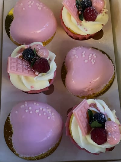The image displays a top-down view of a box containing six pastries. Three of the pastries are heart-shaped and covered in a smooth pink glaze, adorned with small white sprinkles. The other three pastries are cupcakes with swirls of white frosting, topped with a raspberry, a blackberry, a pink chocolate bar, and pink sprinkles. The box appears to be a light brown cardboard, and the pastries are resting on gold or light brown circular bases. The lighting suggests an indoor setting, possibly a bakery or a home. There are no people or animals visible in the image. The image does not contain any information or context to suggest a specific location in Kobeliaky, Ukraine.