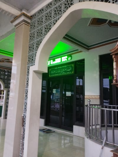 The image captures the entrance area of a mosque in Kecamatan Panji, Indonesia, likely during the late afternoon or early evening. The view is from beneath a large, light-colored archway, looking towards the main entry doors.
A prominent architectural column stands on the left, featuring a light-beige finish and decorative, intricately patterned light-gray tiles with geometric or floral motifs. These patterned tiles also adorn the wall above the column. The ceiling of the arched area and the immediate overhead section are light-colored, illuminated by a vibrant green light source, casting a noticeable green hue on some surfaces.
At the center of the frame are glass double doors, serving as the main entrance. Above these doors, a glowing green digital display shows "17.02 WIB" (5:02 PM Indonesian Western Standard Time) and "SENIN 20 MARET 2023 17" (Monday, March 20, 2023, 5 PM). Below this display, a green sign with a gold border prominently displays the text "DOA MASUK MASJID" (Prayer for entering the mosque). Beneath this title, a prayer in Arabic script is visible, followed by its transliteration/translation in smaller Indonesian text: "Allahummaftahli Abwaba Rahmatika" (O Allah, open for me the gates of Your mercy). The abbreviation "HR Muslim" is also faintly visible, indicating the Hadith source.
To the right of the doors, a polished stainless steel railing with vertical bars defines a curved section, possibly an elevated landing or steps. The floor in the foreground is tiled with a glossy, light-colored material, reflecting the ambient light. A small doormat with striped patterns rests directly in front of the glass doors. No people are visible, and no specific event is taking place, presenting a tranquil scene of the mosque's entrance.