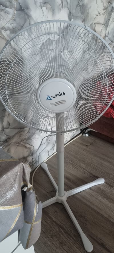 The image shows a close-up, slightly angled view of a white electric fan. The fan has a circular grille with a white circular emblem in the center. The emblem has blue lettering that appears to read "AYAKS" and a stylized blue triangle next to it, along with three vertical lines. The fan's blades are blurred, suggesting it is in operation.
The fan is supported by a white, multi-pronged base and a vertical pole. The pole has textured sections, likely for height adjustment.
The background is a mix of flooring. To the left, there is a light-colored floor with a marble-like pattern in shades of grey and white. To the right, there is a wooden floor with planks oriented horizontally.
A white power cord for the fan is visible on the floor. Partially visible in the foreground and to the right is a folded piece of fabric, possibly a blanket or throw, with a geometric pattern of grey and gold lines. In the upper left background, a dark reddish-brown object with horizontal stripes can be seen, and a glimpse of a metallic object, possibly a lamp base, is also present.
The lighting in the image suggests an indoor setting, possibly during daylight hours, given the ambient light. There are no people visible in the image. The scene depicts a domestic or office environment where a fan is being used.
No specific time of day or weather conditions can be determined from the image. The location context of Kalinkavichy, Belarus, is provided externally.