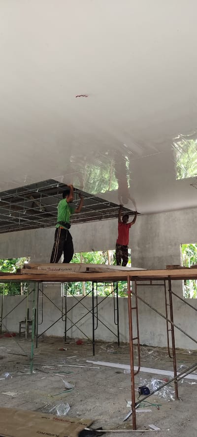 In Langnibong, Indonesia, two construction workers are installing a white, glossy ceiling panel onto a metal grid system suspended from an unfinished concrete structure. The worker on the left, wearing a green shirt and dark pants with striped accents, stands on scaffolding and holds the ceiling panel in place while working. The worker on the right, in a red shirt and patterned pants, also stands on scaffolding and is in a similar posture, holding a section of the ceiling. The ceiling material appears to be made of PVC, as indicated by a partially visible cardboard box on the scaffolding that reads "PLAFON PVC". The background reveals an open construction site with more scaffolding, concrete walls with openings looking out to foliage, and a dusty, debris-strewn floor with construction materials scattered around. A coiled red and black wire hangs from the ceiling near the top center of the image.