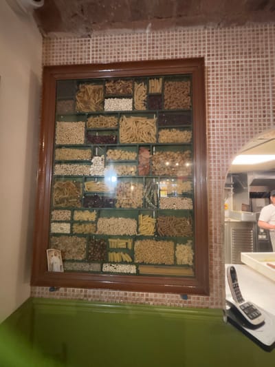 The image shows a wall inside a restaurant in Bradford, United Kingdom. It is located at approximately 53.794° N, 1.752° W. The wall is decorated with a large wooden frame, containing a glass display case filled with a variety of dried pasta and beans, arranged in a grid pattern. There are 36 individual compartments. Each compartment is filled with a different kind of pasta or beans. The wall is painted green, with a small section of red tiled trim at the base of the wall. There is a white phone on a white surface to the right of the frame. The phone cord is visible under the phone. Through an arched opening behind the wall, one can see the edge of a stainless-steel kitchen and a person wearing a white shirt and a black apron working at a counter. The person's head and shoulders are obscured by the opening.