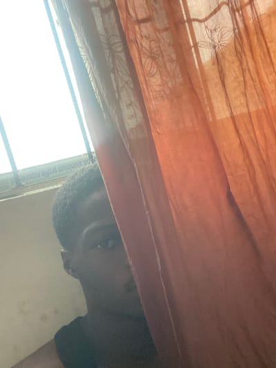 The scene is an interior location in Igbessa, Nigeria. A dark-skinned individual with short dark hair is partially visible, positioned behind an orange-brown sheer curtain. Only the left side of the individual's face, including the left eye, is discernible in the left-central portion of the image, oriented towards the camera. A dark garment covers the visible left shoulder. The curtain, which features a subtle, translucent pattern, extends from the right edge of the frame to approximately the center, partially obscuring the individual. In the upper left quadrant, a window with a white aluminum frame is visible, through which bright natural light enters, backlighting the scene. Inverted white text, specifically "aluminium profile expone", is legible on the window frame. A plain light-colored wall constitutes the background on the far left of the image.