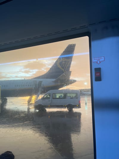 The image is taken from inside a vehicle, looking through a rain-streaked window. Outside, an airplane is visible, specifically its tail fin, which features the Ryanair livery. A white service van is also present. The presence of an airplane suggests the location is an airport. The text on the emergency call button is difficult to read, but the presence of Ryanair points to the possibility of Alicante–Elche Miguel Hernández Airport, located in the city of Elx / Elche, Spain. The light suggests either sunrise or sunset. There are no visible people in the image aside from a possible head at the very bottom-right.