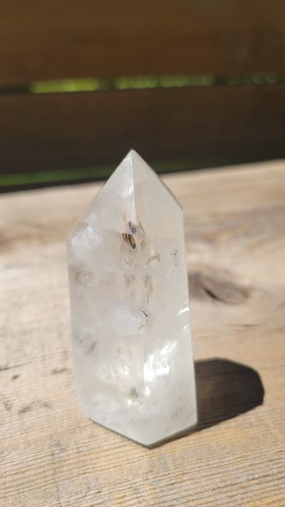 A clear quartz crystal point is shown standing upright on a weathered wooden surface, likely a table or deck. The crystal is elongated with a hexagonal base and a pointed apex. Inside the crystal, there are visible inclusions and fractures, giving it a natural, imperfect appearance. The lighting suggests an outdoor setting, with a shadow cast by the crystal onto the wooden surface. In the background, out of focus, are horizontal wooden planks with blurred green areas between them, possibly indicating a railing or fence with vegetation beyond. The scene appears to be a still life composition, with no people or animals present. The location is not specified in the image or any accompanying metadata.