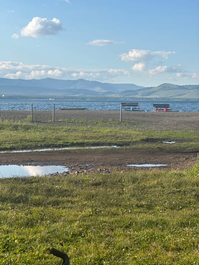 This is an outdoor, eye-level, full shot image of a lake and its surroundings. The sky is a light blue with scattered white clouds, some of which are large and puffy. In the middle ground, a body of water with choppy waves extends across the frame, meeting a pebbly shore. Two docks or platforms with benches are visible on the water. The foreground is dominated by green grass, with a muddy path and puddles of water closer to the shore. In the far distance, rolling hills with a faint green hue are visible under the hazy sky. The scene appears to be located near the city of Якты-Куль, Russia, possibly a recreational area by a lake.