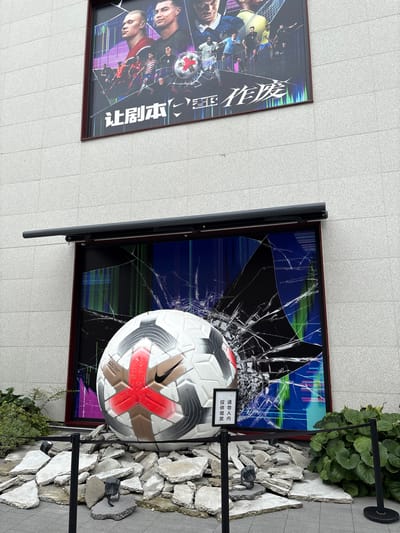 A large, cracked digital screen displays a dynamic image of soccer players and a soccer ball with a red and gold pattern and a Nike logo. The screen is mounted on a gray, textured building. In front of the screen, a giant soccer ball, identical to the one on the screen, rests on a pile of broken concrete slabs. Black ropes and stanchions, along with two spotlights, are placed around the display. A small sign with Chinese characters is visible near the base of the soccer ball. The location is in the city of Qingpu District, China.