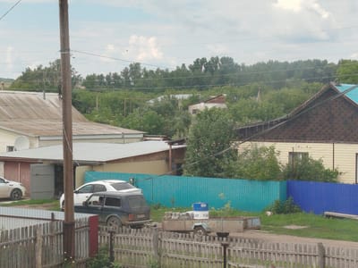 The image depicts a street scene in the village of Ziyanchurino, Russia. In the foreground, a wooden fence runs horizontally across the frame, with a wooden utility pole situated to the left of the center. Behind the fence, a dark blue Lada Niva is parked, facing to the right. To the left of the Lada, a white sedan is partially visible, parked in front of a garage. A trailer with several cylindrical objects and a larger white and blue barrel is positioned to the right of the Lada. The background features a turquoise and blue corrugated metal fence, separating the street from a residential area. Beyond the fence, various buildings with sloping roofs are visible, interspersed with lush green trees and foliage. The sky is overcast with scattered clouds. The overall impression is that of a typical rural or suburban residential area.