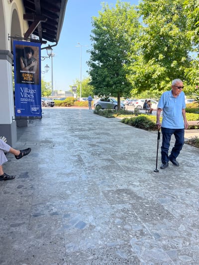 An elderly Caucasian man, wearing a light blue polo shirt and dark blue jeans, walks on a stone-paved walkway with a cane in his right hand. He is looking down and to his right. He wears dark-rimmed sunglasses and has short gray hair. Behind him, a banner advertising "Village Vibes" hangs from a building. The banner features text in Italian and an image of a person. Trees with green foliage are visible in the background, along with a parking lot and a clear blue sky. In the lower left corner of the image, the legs and feet of another person are visible, seated on a bench. This person is wearing light gray pants and black sandals. The scene appears to be an outdoor public area, possibly a shopping center or a pedestrian walkway, in Rodengo, Italy.