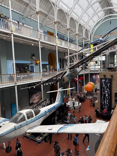The image shows the Grand Gallery of the National Museum of Scotland in Edinburgh, United Kingdom. The vast, multi-story space features a distinctive glass and iron roof structure. Suspended from the ceiling are several aircraft, including a light aircraft in the foreground with the registration G-BGXB and the text "Piper Tomahawk" visible. Above and behind it, another aircraft, possibly a glider, is also suspended. The museum floor is a red tiled surface with a geometric pattern. Visitors are seen on various levels of the gallery, observing the exhibits. Some are standing and looking up at the aircraft, while others are seated or walking around the displays on the ground floor. The walls of the museum are painted in a muted blue, and the upper levels have white balustrades. Various exhibits and displays are visible on the walls and in display cases on the lower levels, including what appears to be a collection of photographs or postcards in a tall, black frame on the right side of the image. The overall scene depicts a bustling museum environment with impressive aeronautical exhibits.