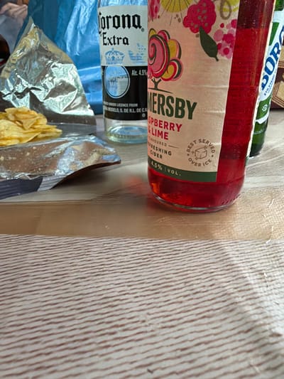 The image depicts a close-up view of a table surface with several items, suggesting a casual gathering or snack time. Dominating the foreground is a glass bottle of Sommersby Raspberry & Lime flavored cider, its contents a vibrant red and the bottle exhibiting condensation. To the left of the cider bottle is a partially opened bag of potato chips, with a few chips scattered on the surface and a crumpled silver foil wrapper visible. Behind these items, a bottle of Corona Extra beer is partially visible, its label showing the iconic crown logo and "Extra." A blue plastic bag is also present in the background. The surface on which these objects rest appears to be a table with a light-colored wood grain texture. The lighting suggests an indoor setting, possibly during the day. The scene is devoid of any human presence or activity. The overall impression is one of casual refreshment and snacking, with no clear indication of the specific location beyond being an indoor table setting. There is no information in the image that suggests the location is the city of Pravets, Bulgaria.