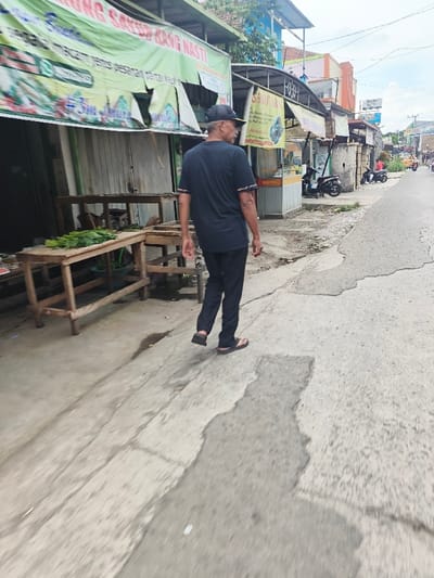 An older man in a dark t-shirt, dark pants, a baseball cap, and sandals walks on a cracked concrete street. To his left are a couple of empty wooden tables next to a storefront with a weathered green sign. Other shops line the street in the background, along with parked motorcycles. A street stretches into the distance, where faint figures suggest the presence of people and vehicles. The sky above is overcast.