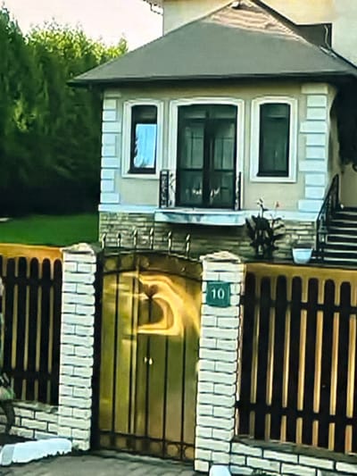 The image shows the exterior of a residential property, likely a house, situated in the city of Dondușeni, Moldova. In the foreground, there is a dark wooden fence with vertical planks. The fence is bordered by white brick pillars, one of which has a green house number sign displaying "10". Behind the fence, a golden gate is visible. The property appears to be well-maintained, with a manicured lawn and trees in the background. The house itself has a light-colored facade with stone accents and a dark roof. There are windows and a balcony on the upper level. The overall scene suggests a peaceful, suburban setting.