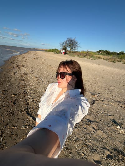 The media file depicts a selfie taken by a person on a sandy beach near Port-Katon, Russia. The subject is wearing sunglasses and a white shirt with a pattern of small red and yellow hearts. The background shows a clear blue sky, a shoreline with gentle waves, and a vegetated area with a bare tree and some shrubs. In the distance, a few other people are visible near the vegetation, some of whom appear to be seated. The sand on the beach is dark and contains small pebbles and debris. The lighting suggests it is daytime, likely late afternoon given the shadows. The overall mood appears relaxed and leisurely, consistent with a day at the beach. There is no visible text in the image.