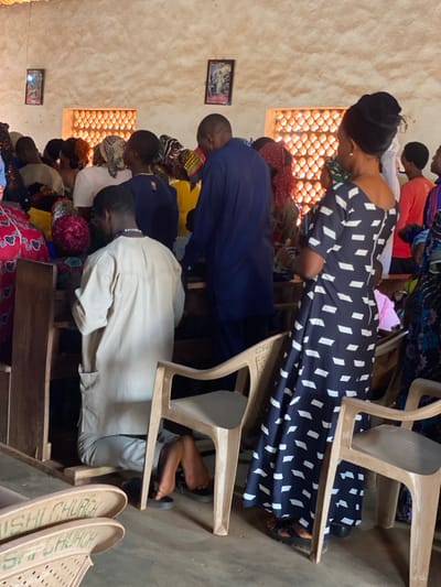 The image depicts an indoor gathering within a "SHI CHURCH" in Nev, Nigeria. Approximately 15-20 individuals are present, primarily viewed from the rear or side, participating in a communal event. In the foreground left, a male subject kneels on the floor, facing away from the camera, wearing a light-colored tunic and trousers. His feet, clad in black sandals, are visible beneath a wooden pew. To the right, a female subject stands, partially turned away, dressed in a dark blue patterned gown with white parallelogram shapes and a dark headwrap. Multiple light-colored plastic chairs are interspersed among dark wooden pews. A plastic chair in the foreground displays "GWO SHI CHURCH" embossed on its backrest. The light brown, textured walls feature two rectangular window openings fitted with orange-brown terracotta lattice blocks, allowing natural light penetration. Two framed religious pictures are mounted on the wall between the windows. The floor surface is a uniform grey-brown.